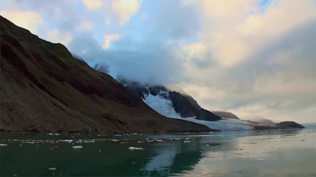 Clouds linger over rugged landscape as ice meets water. Small pieces of ice float along surface, reflecting sky near Svalbard glacier. Mountains reach toward heavens in this scenic arctic landscape.