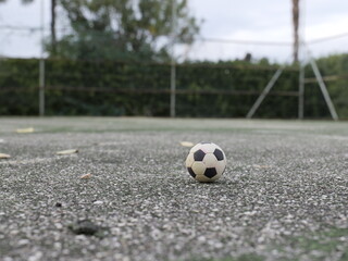 Soccer ball on empty outdoor court ground level view. Sports field with fence and trees in background. Urban recreational area for football training and games.