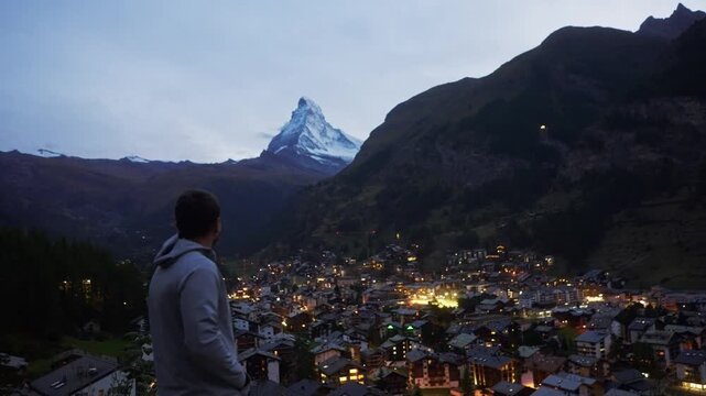 Traveler looking at Zermatt town and Matterhorn mountain during blue hour. Scenic view of glowing city lights in Swiss Alps, Switzerland