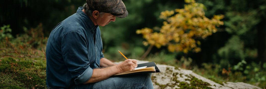 A thoughtful man is seated on a rock in nature, writing notes in his notebook with a pencil, surrounded by greenery and autumn leaves, showcasing tranquility and reflection. - Powered by Adobe