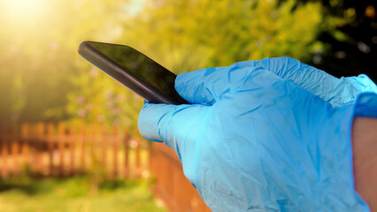 Hands in blue gloves holding a smartphone outdoors in garden. Man using a phone.