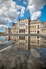 Naklejka premium Budapest, Hungary - July 27, 2025: Reflection of the Hungarian Parliament Building in the shallow pool at Kossuth Lajos Square under a dramatic summer sky 