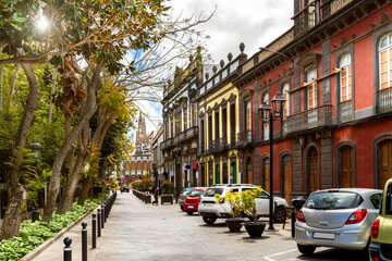 Sunny old town street in Arucas with colorful facades, trees, planters and distant cathedral view