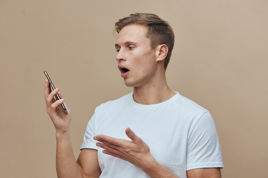 surprised young caucasian man wearing white t-shirt hold smartphone use voice assistant talk on beige background digital lifestyle concept