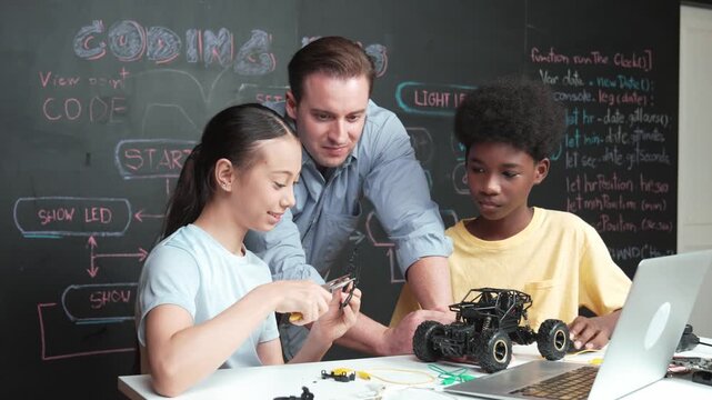 Caucasian teacher pointing at laptop screen while explain car model system at blackboard with coding or programming code written. Happy diverse student learning about robotic construction. Edification