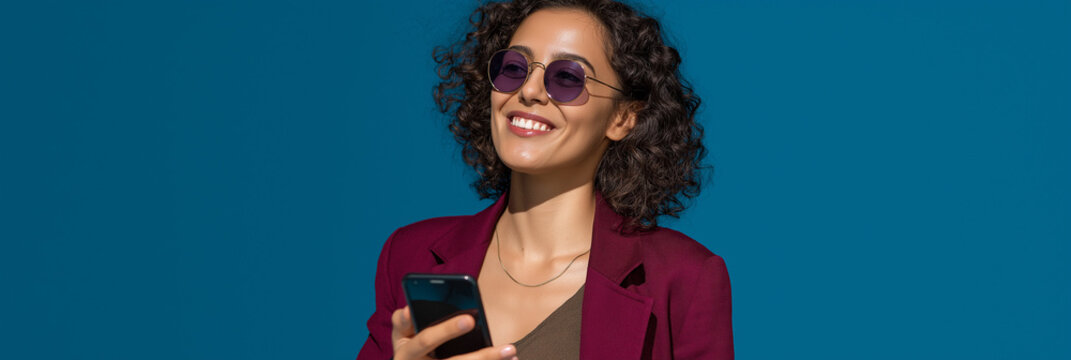 A joyful woman stands outdoors under a clear blue sky, smiling brightly while using her smartphone, capturing a moment of happiness and connection in a vibrant atmosphere.