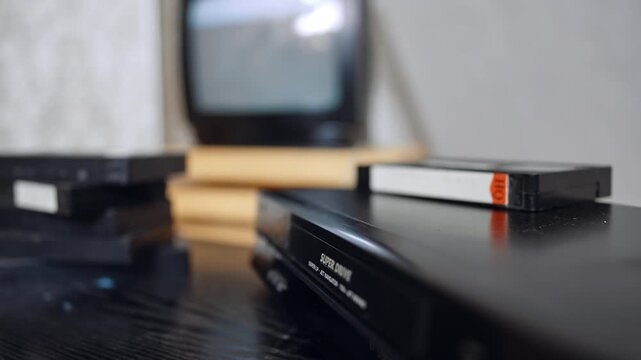 VHS player, several tapes and shimmering screen. A cassette appears from the player and hand puts it on the pile.