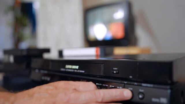 Old Caucasian man's hand pressing a button on the retro recorder. Screen shimmering at blurred backdrop. A cassette appears and male puts it on the pile.