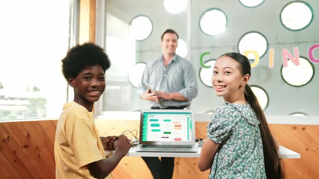 Diverse pupil study programming system while turn around to look at camera. Caucasian teacher standing and teaching multicultural student about coding prompt at STEM technology class. Edification