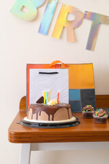 asty chocolate birthday cake with frosting and a candle, accompanied by colorful cupcakes and gift bags on a wooden desk, ready for a home office celebration.