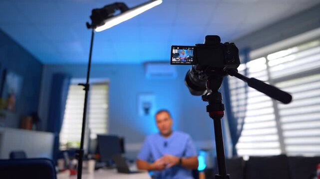 Light and camera directed on the man at blurred backdrop. Male in blue uniform talks gesturing in front of camera for his blog. Selective focus.