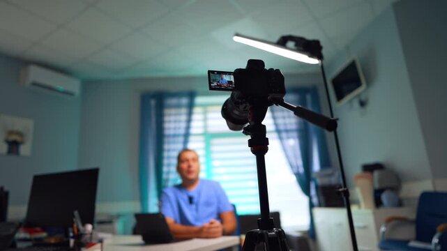 Modern camera on tripod recording a man talking at backdrop. Low angle view. Video blog creation. Selective focus.