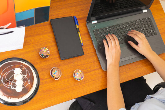 Close-up of hands typing on a laptop keyboard next to three colorful birthday cupcakes with frosting and sprinkles. Concept of remote celebration, online communication, and modern lifestyle combining 
