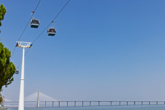Cable cars against a clear blue sky over the Tagus River in Lisbon's Parque das Na&ccedil;&otilde;es, with the expansive Vasco da Gama Bridge in the distance