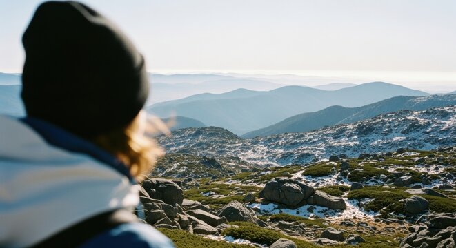 Female hiker enjoying a mountain summit view. Expansive winter landscape with snowy peaks and rocky terrain. Adventure travel and outdoor exploration lifestyle