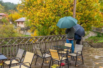 Couple with Umbrellas Standing on a Wet Stone Patio of a Traditional Cafe in Vovousa Village, Pindos, Surrounded by Autumn Foliage