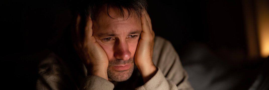 A somber man with his hands on his head, displaying feelings of stress and contemplation against a dark background, symbolizing the weight of thoughts and worry.