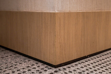 Close-up of a sharp corner where light wood paneling meets a dark base and a patterned beige and brown carpet. Interior material detail