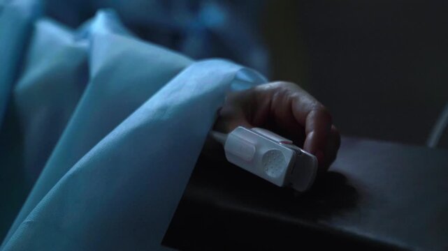 Close up of an unconscious patient's hand with a pulse oximeter on the finger, lying still in a dark hospital room, symbolizing severe illness, intensive care or the end of life