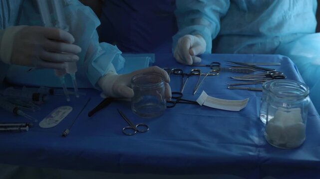 Professional surgeon and assistant preparing biomaterial for a plastic surgery operation, with sterile instruments like syringes, scissors, and forceps laid out on a blue medical drape