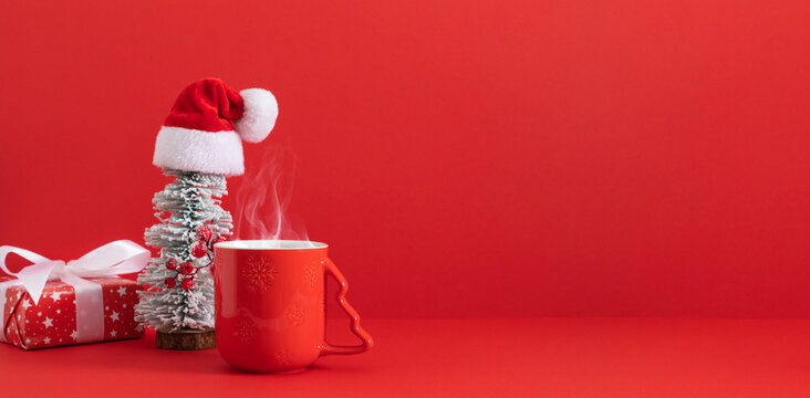A cozy Christmas arrangement featuring a steaming red mug and a festive gift box with a miniature Christmas tree topped with a Santa hat, on a vibrant red background with room for text.