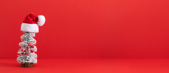 Festive Christmas minimalism. A small Christmas tree topped with a tiny Santa hat on a solid red background with plenty of room for your holiday message. Banner