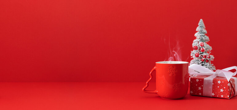 Cozy Christmas winter morning: a steaming red mug next to a festive gift box and a miniature Christmas tree on a bright red background with space for text. Banner