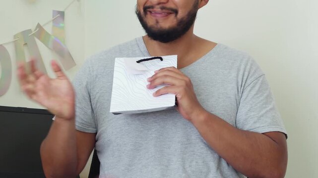 Happy man with party glasses enjoys his birthday while holding a colorful present against a festive backdrop.