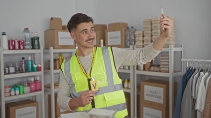 Young hispanic man wearing reflective vest in charity donations center taking a video call surrounded by packed goods showcasing his volunteer work.