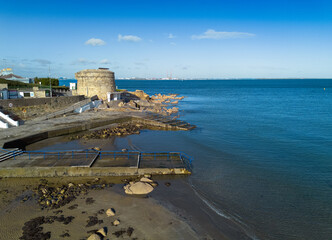 A beach with a dock and Martello Tower Number 14, Seapoint, Blackrock, Dublin. The water is calm and the sky is clear.
