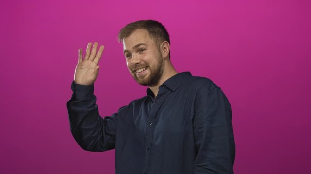 Man waving right hand in studio wearing navy shirt against vibrant pink studio wall; friendly greeting.