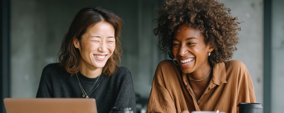 Two women coworking with laptops and coffee cups, smiling and collaborating in a modern office