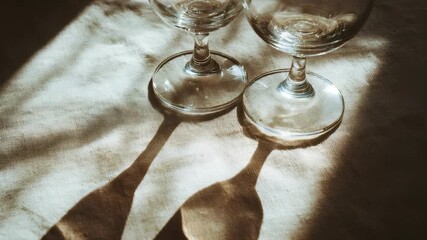 Close up of two clear wine glasses with artistic shadows on a neutral tablecloth, illuminated by golden sunlight. The scene suggests intimacy, simplicity, and modern lifestyle aesthetics