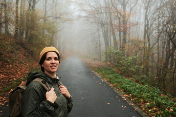 Smiling woman with backpack stands on quiet foggy road in autumn forest. Calm travel and exploration concept