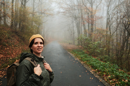 Smiling woman with backpack stands on quiet foggy road in autumn forest. Calm travel and exploration concept