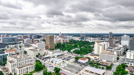 Panoramic aerial view of Austin, Texas skyline under dramatic cloudy skies. The Texas State Capitol building is prominent amidst a dense urban landscape with skyscrapers and roadways. A moody