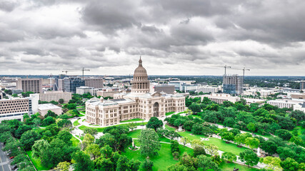 Dramatic aerial view of Texas State Capitol building amidst a cloudy Austin skyline. The iconic dome dominates the landscape, framed by modern skyscrapers and construction cranes under a moody sky. A