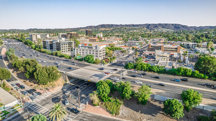 Aerial view of a busy highway cutting through a suburban landscape with buildings and hills in the distance. Cars flow along the road under an overpass on a sunny day. Ideal for business