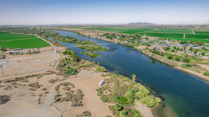 Aerial view of a river landscape in Arizona. Agricultural fields stretch across the horizon under a clear blue sky. A bridge spans the waterway with a small RV visible. Ideal for nature, agriculture