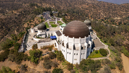 High-angle aerial shot of the Griffith Observatory in Los Angeles. The iconic dome structures are prominently displayed amidst lush greenery and rolling hills. People visible near the observatory.