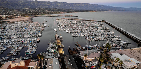 Aerial view of a bustling harbor filled with boats at dusk. Rolling hills in the background under a dusky sky. Reflective water adds to the serene atmosphere.