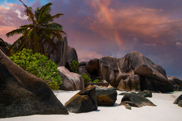 Granite boulders and white sand define Anse Source d Argent on La Digue, Seychelles, as a pastel sunset glows with purples and oranges and a faint rainbow appears.