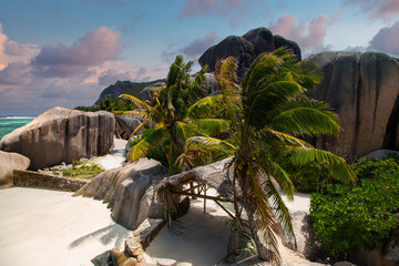 Granite boulders frame a narrow sandy path to turquoise water at left. Coconut palms and a thatched...