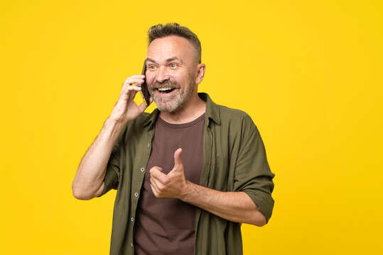 Attractive senior 50 man having joyful happy conversation over smartphone, amazed smilling face, open mouth and hand thumb up gesture. man talking on mobile phone on yellow background