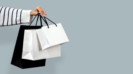 Woman hand in striped sweater holding white and black paper shopping bags, blue background. Concept of e-commerce, online purchases delivery, holiday seasonal discounts.Copy space, sale banner, mockup
