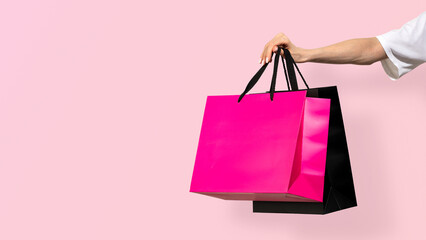 Female hand holding glossy pink and black paper shopping bags on pastel pink background. Concept of shopping, seasonal sales, promotional retail marketing and consumer experience. Copy space, banner