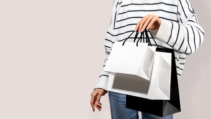 White and black paper shopping bags in woman's hand in striped sweater and jeans on beige neutral background. Concept of lifestyle shopping, clothing seasonal discounts. Copy space, banner, mockup