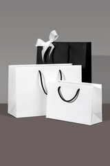 Three paper shopping bags, white and black with handles on grey background. Concept of premium gifting, stylish packaging, holiday shopping display and elegant branding for festive occasions. Vertical