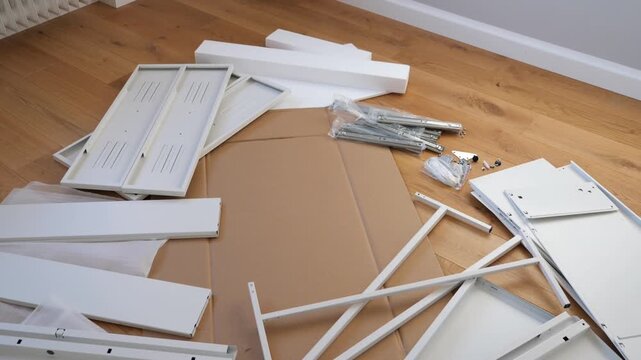 DIY furniture assembly parts laid out on wooden floor &ndash; white panels, metal rods, screws, bolts, and fasteners organized before building shelf, desk, or cabinet.