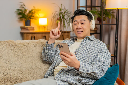 Happy overjoyed middle-aged man in wireless headphones relaxing lying on couch using phone at home choosing, listening favorite energetic disco dancing music. People weekend leisure activities.